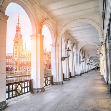 Langer, überdachter Steingang entlang der Alster mit Blick auf das Rathaus während des Sonnenuntergangs.