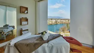 Massageliege mit Handtüchern in einem kleinen Raum mit bodentiefen Fenstern über der Aare.