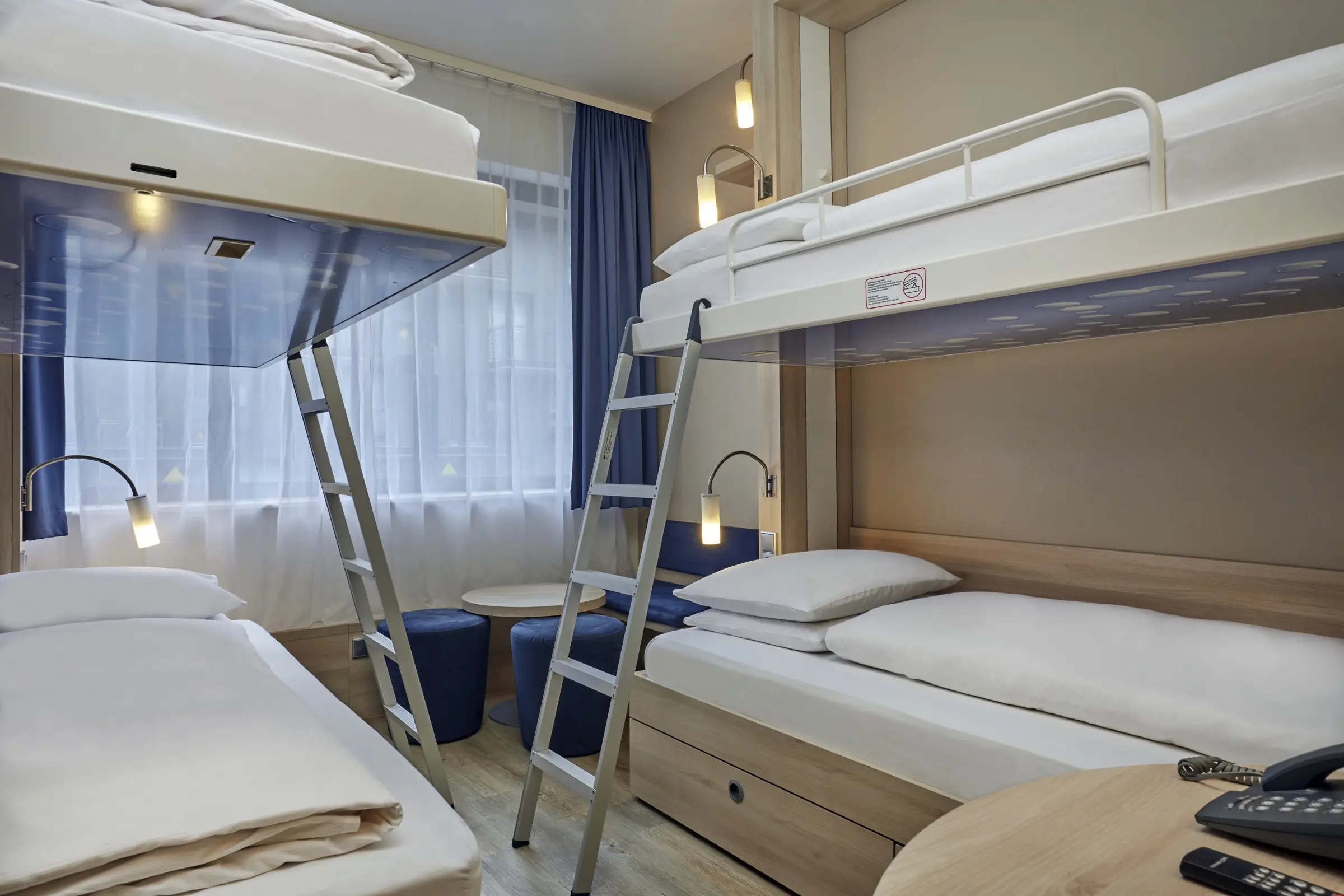 Nahaufnahme von zwei Etagenbetten im Hotelzimmer. Vor dem Fenster steht eine kleine Sitzeecke mit Hockern.