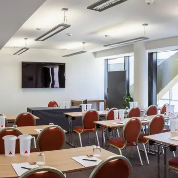 Heller Konferenzraum mit ein paar Sitzreihen. An der Wand hinter dem Rednertisch hängt ein großer Fernseher.