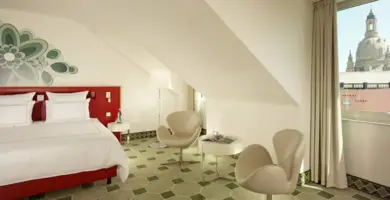 Hotelzimmer mit einem roten Doppelbettauf der linken Seite. Auf der rechten Seite stehen zwei Stühle und ein Tisch.