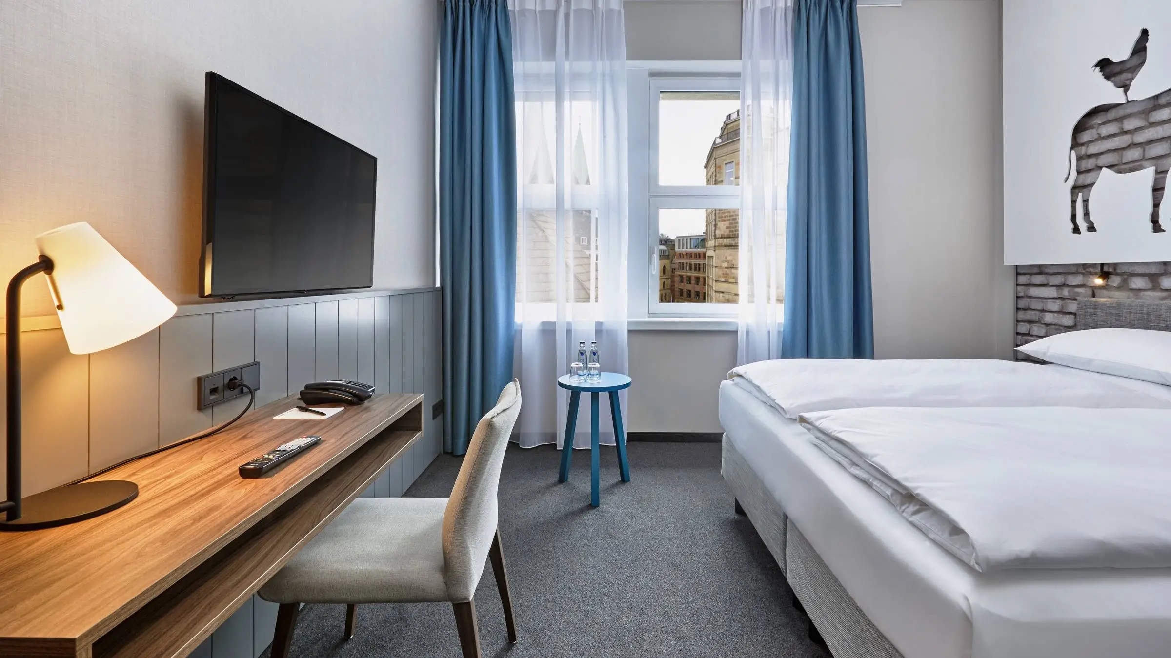 Hotelzimmer mit einem Doppelbett auf der rechten Seite und einem Schreibtisch auf der linken Seite. Über dem Schreibtisch hängt ein Fernseher.