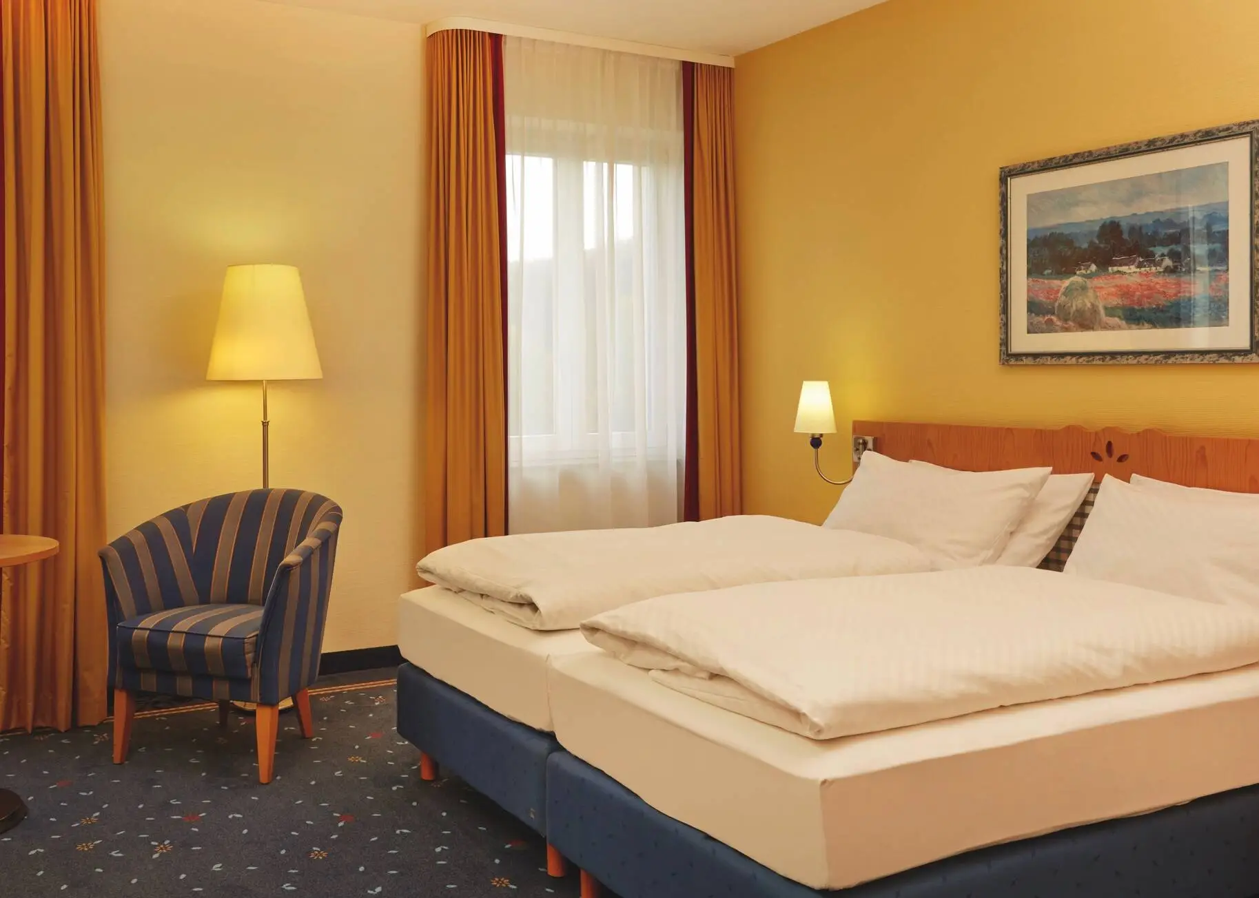 Camera d'albergo illuminata con due letti singoli uniti e una poltrona a righe blu e oro accanto a un tavolo rotondo. Sul tavolo ci sono due bottigliette d'acqua e bicchieri.