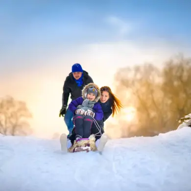 Eine Mutter fährt mit ihrem Kind auf einem Schlitten einen kleinen Hügel hinunter. Der Vater hat sie angeschubst.