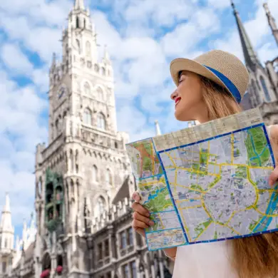 Frau mit Sonnenhut hält einen Stadtplan in der Hand. Sie steht vor dem Münchner Rathaus und schaut es dabei an.