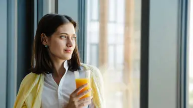 Frau im Businessoutfit sitzt lächelnd mit einem Glas Orangensaft am Fenster.