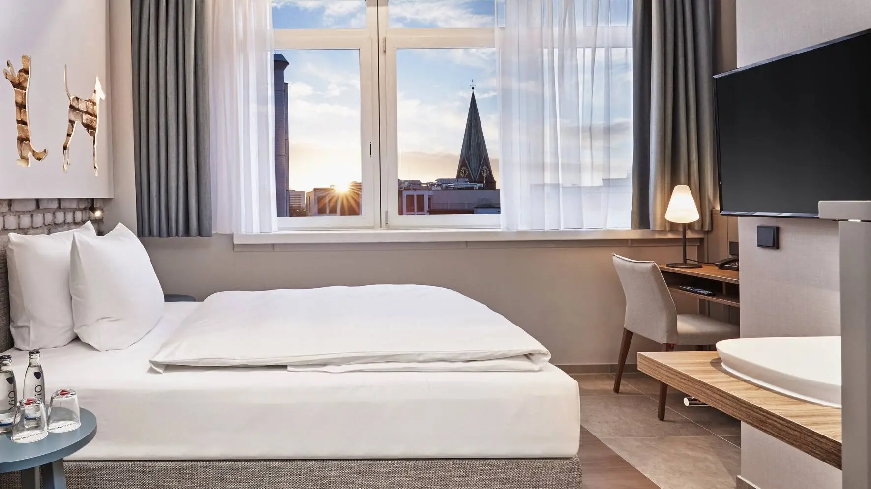 Chambre d'hôtel avec un lit et un bureau devant. Au premier plan, sur la gauche, se trouve une armoire avec une machine à café et sur la droite, une télévision est suspendue.