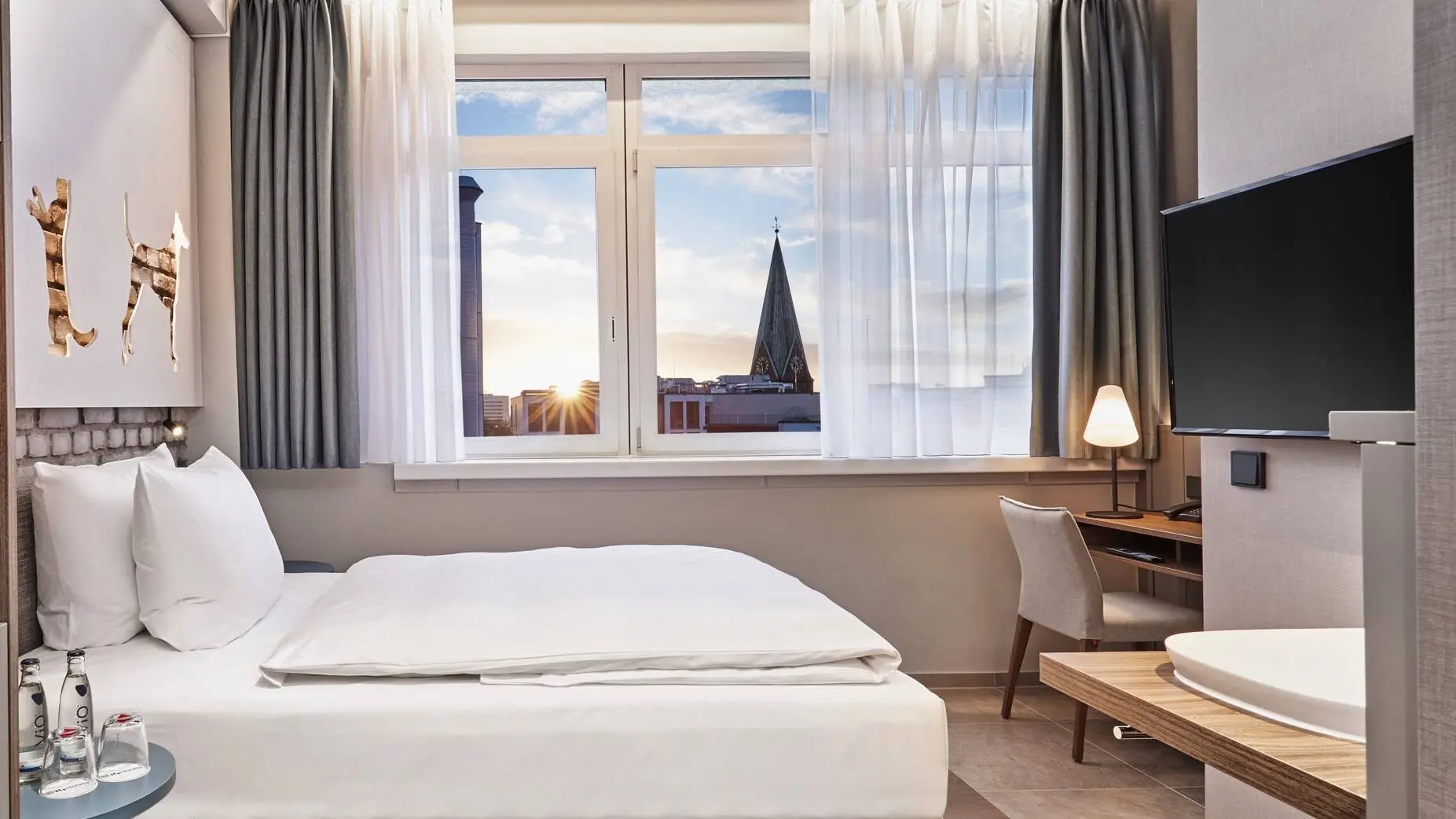Hotelzimmer mit einem Bett und einem Schreibtisch davor. Im Vordergrund steht auf der linken Seite ein Schrank mit einer Kaffeemaschine und auf der rechten Seite hängt ein Fernseher.