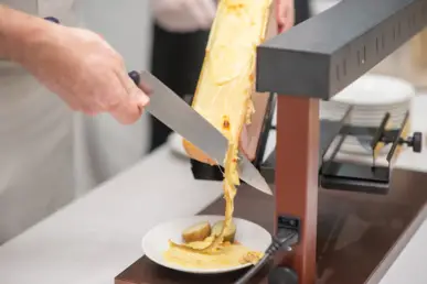 A cook cuts melted cheese from a cheese wheel for raclette with a knife.