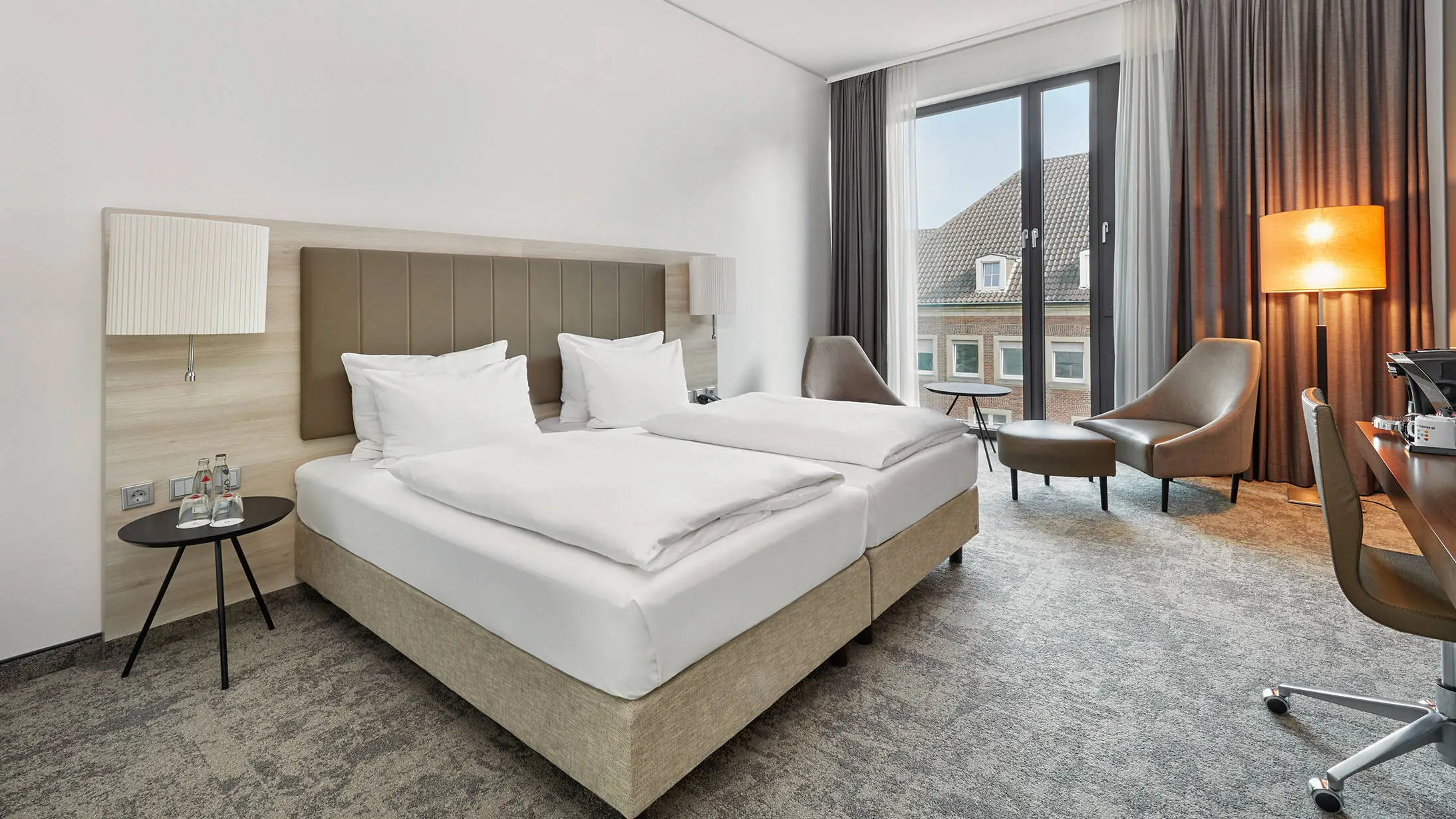 Doppelbett im Hotelzimmer mit einer Sitzecke und einer Lampe vor dem Fenster.