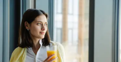 Frau im Businessoutfit sitzt lächelnd mit einem Glas Orangensaft am Fenster.