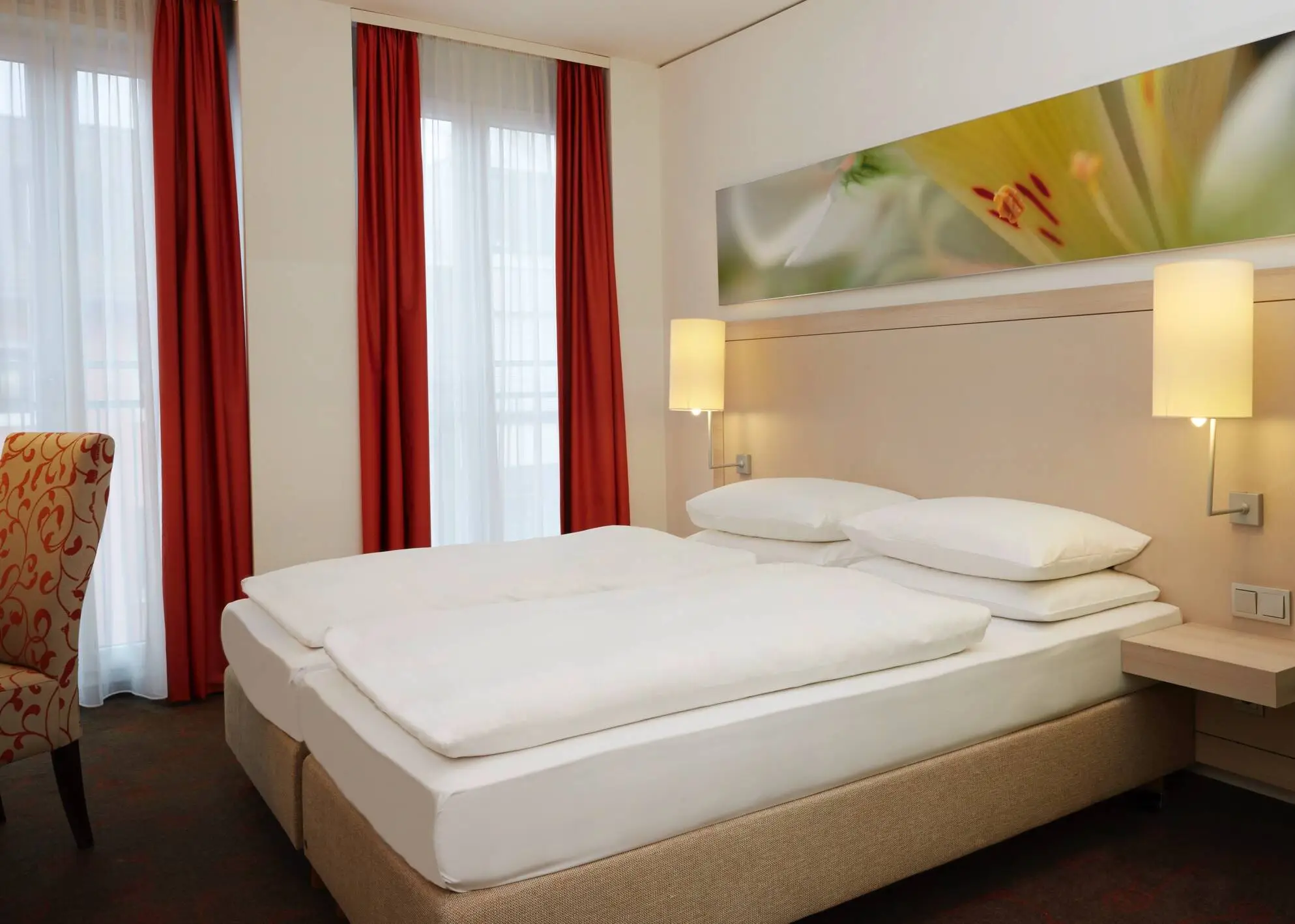 Großes Doppelbett im Hotelzimmer. Am Bett hängen zwei kleine Lampen.