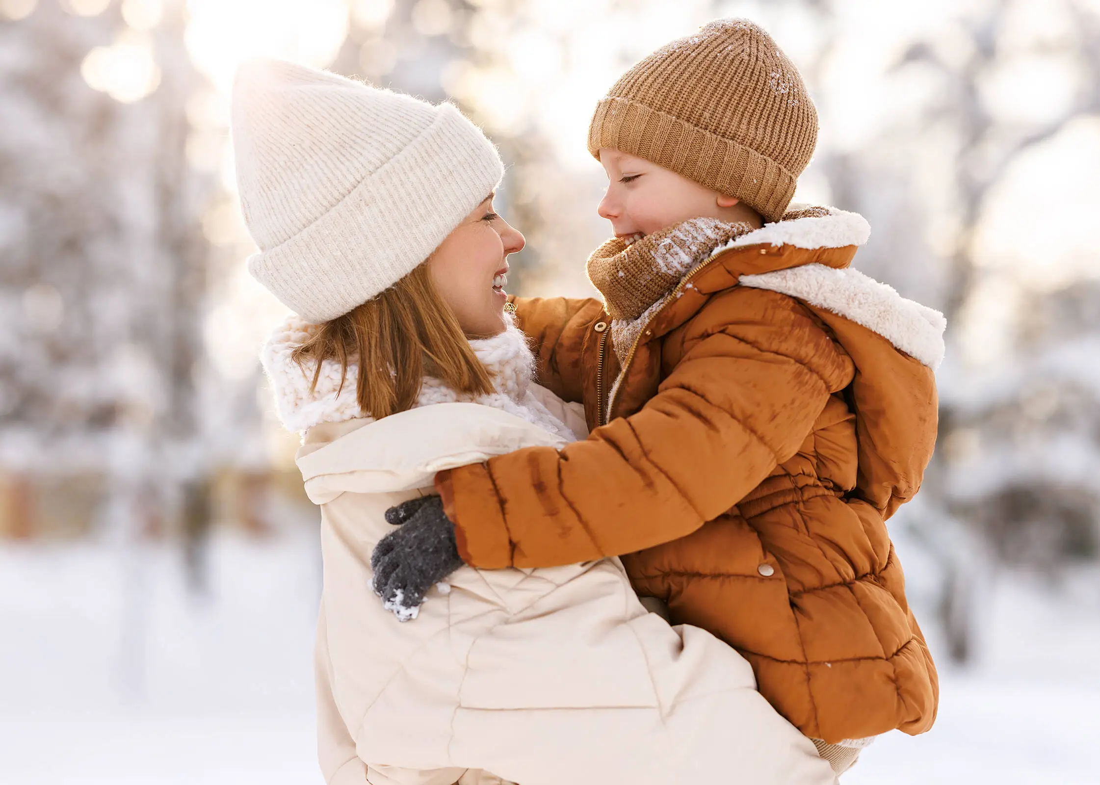 Une femme  vêtue d'une veste blanche et d'un bonnet tient dans ses bras un enfant vêtu d'une veste et d'un bonnet marron. Il y a beaucoup de neige en arrière-plan.