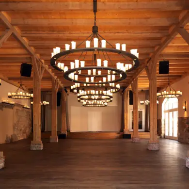 Großer Tagungsraum mit  Holzstützen und großen Kerzenleuchtern an der Decke. Die Wand ist zum Teil gemauert und im Hintergrund ist eine Bühne.