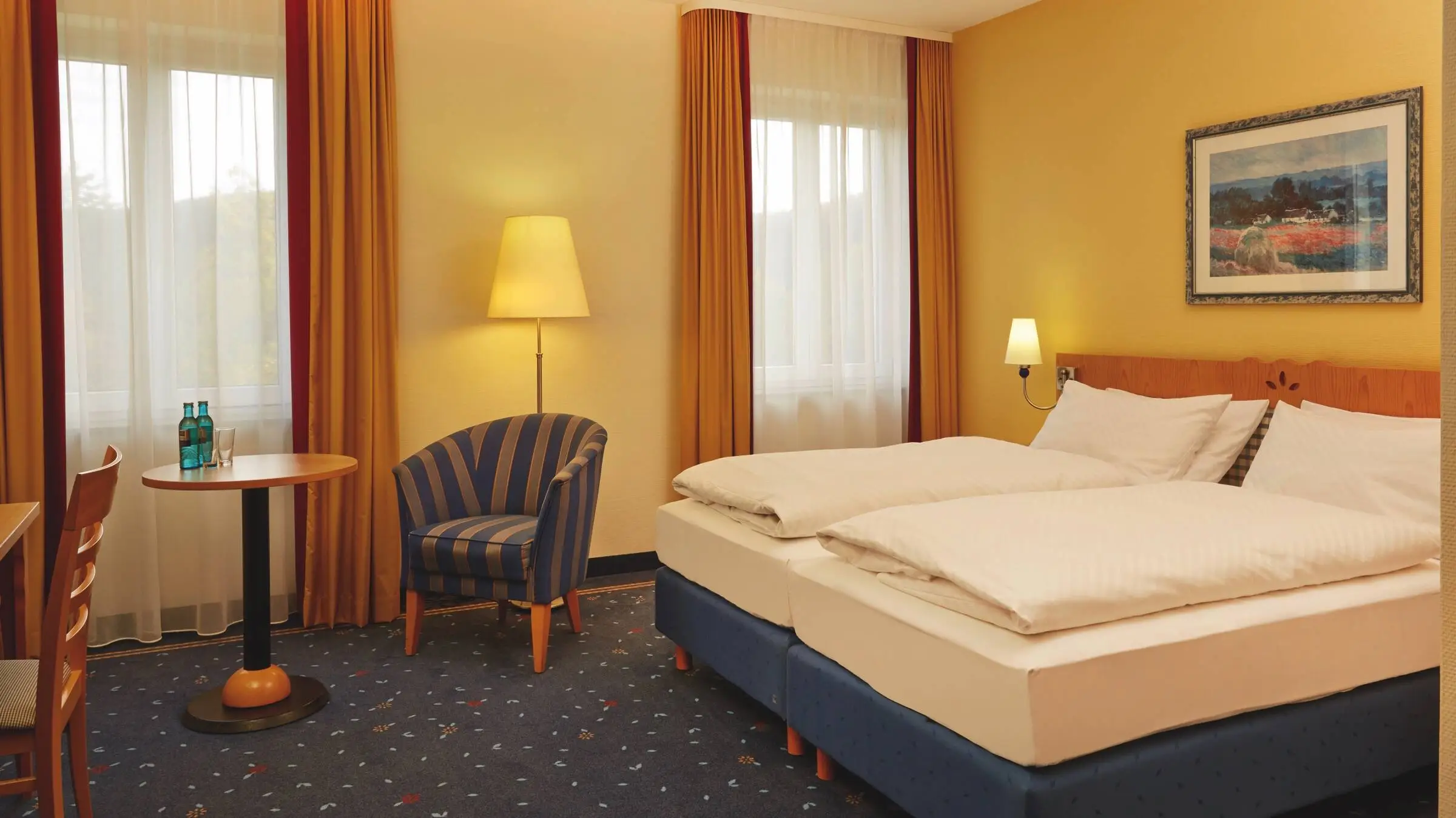 Habitación de hotel iluminada con dos camas individuales juntas y un sillón a rayas azules y doradas junto a una mesa redonda. Sobre la mesa hay dos botellas pequeñas de agua y vasos.
