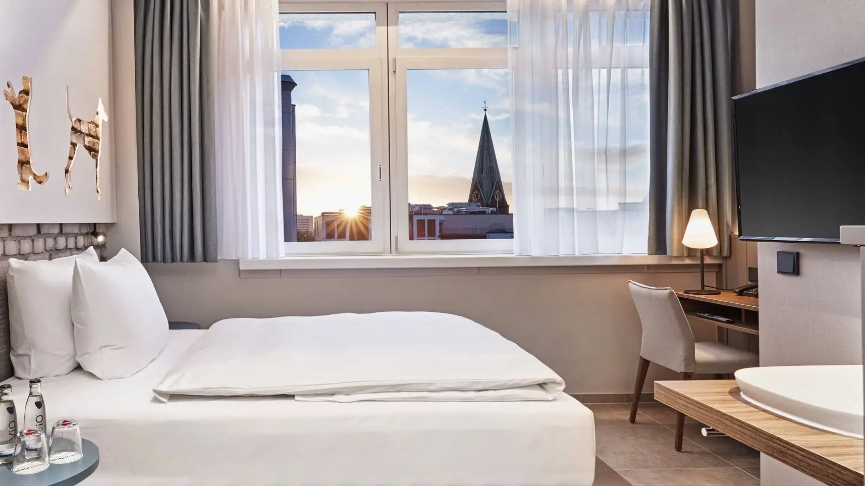 Hotel room with a bed and a desk in front of it. In the foreground on the left is a cupboard with a coffee machine and on the right hangs a television.