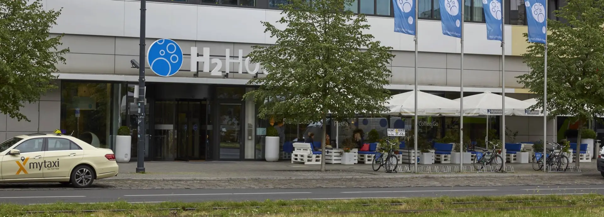 Blick über die Straße auf den Eingangsbereich vom Hotel. Neben dem Eingang stehen Sitzgruppen mit großen Sonnenschirmen.