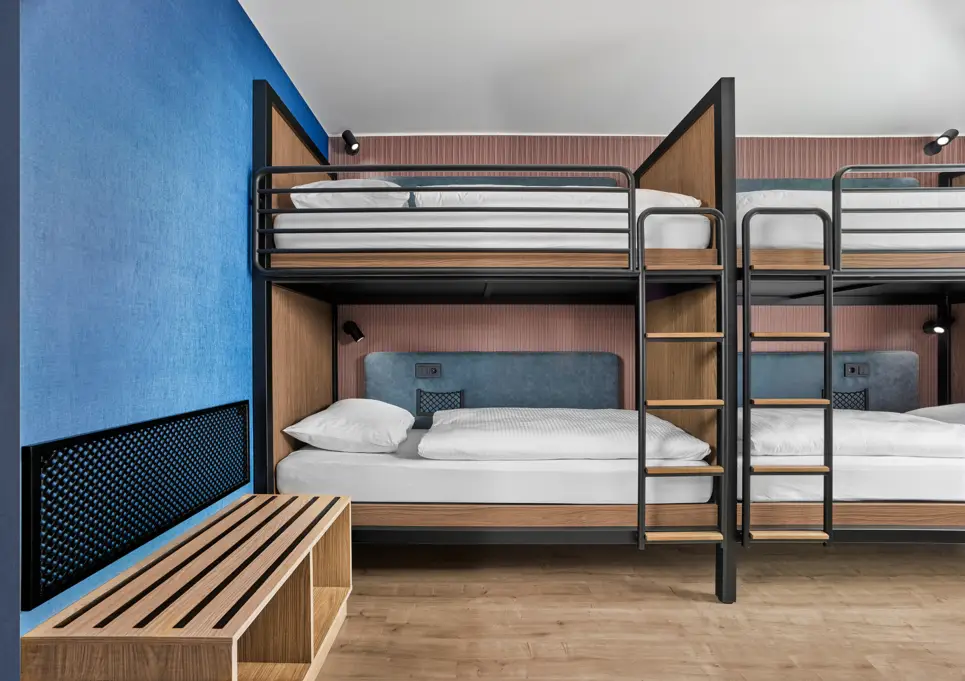 View of bunk beds with ladders. To the left in front of the beds is a wooden frame for sitting or storing bags.