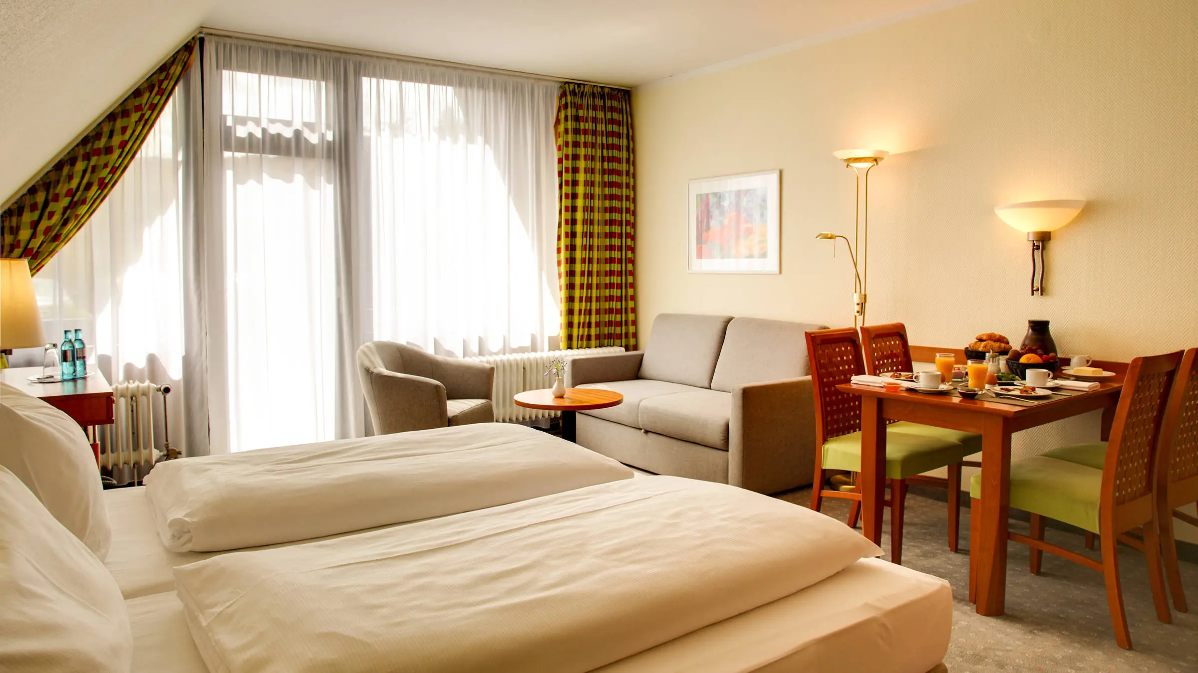 Großes Hotelzimmer mit einem Doppelbett und einem großen Fenster. Vor dem Bett steht ein Esstisch und eine kleine Sitzecke mit einer Couch.