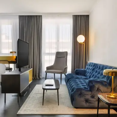 Hotelzimmer mit einer großen, dunkelblauen Samtsofa. Vor dem Sofa steht ein Schwarzgoldener Fernsehschrank mit einem Flachbildfernseher darauf.