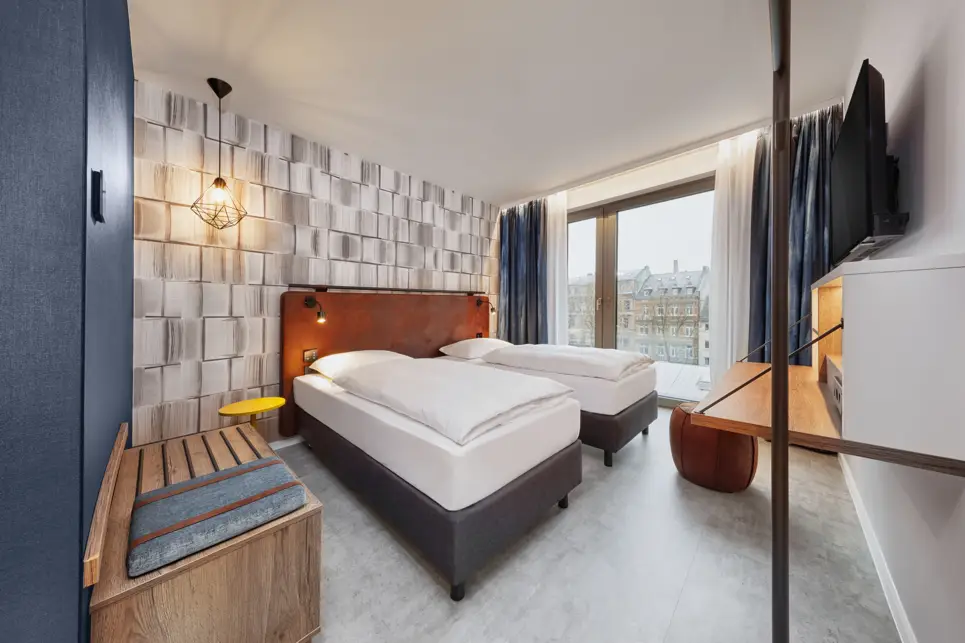 Doppelbett in einem Hotelzimmer mit einer Ablagebank auf einer Seite und einem Schreibtisch auf der anderen Seite.