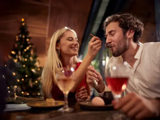 <span>Klassisches 4-Gänge-Menü</span><span> im Familienstil</span> Eine Frau füttert einen Mann mit einem goldenen Löffel. Im Hintergrund steht ein Weihnachtsbaum.