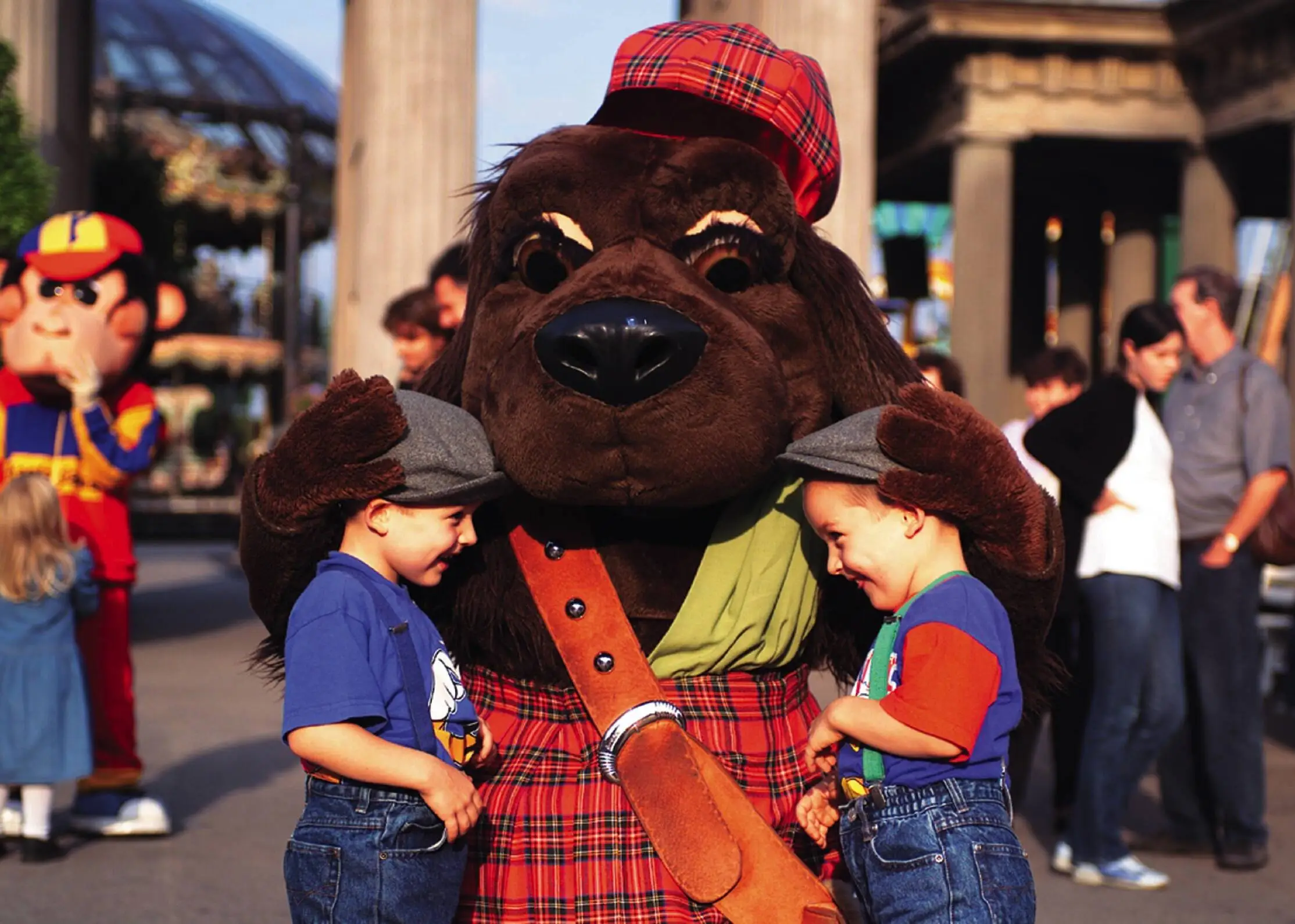 Two small children stand happily in front of a dog mascot. The two children look at each other, the mascot has both paws on the children's heads.
