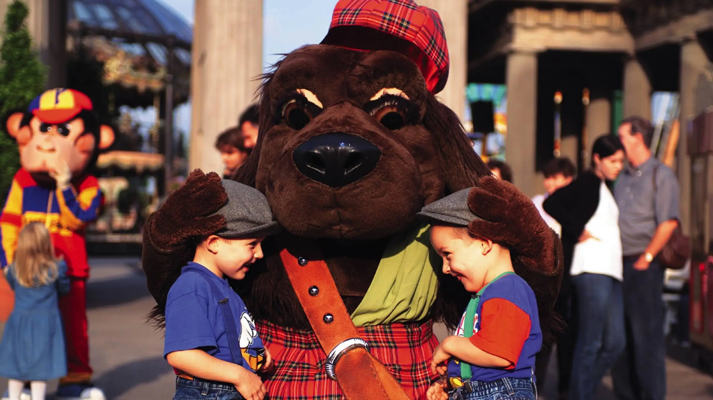 Zwei kleine Kinder stehen glücklich vor einem Hunde-Maskottchen. Die beiden Kinder shcauen sich an, das Maskottchen hat beide Pfoten auf den Köpfen der Kinder.