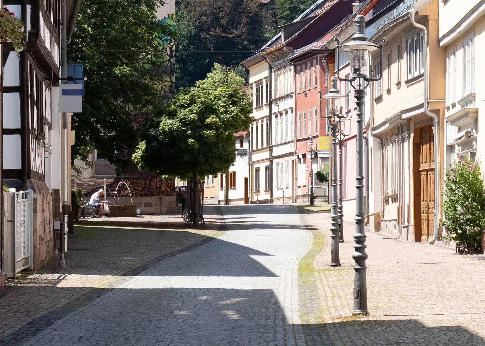 Idyllische Straße in der sommerlichen Altstadt von Friedrichroda mit Fachwerkhäusern und Laternen am Straßenrand. Im Hintergrund Bäume und ein Mann, der auf einer Bank neben einem Brunnen sitzt.