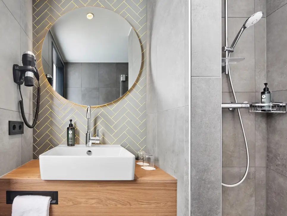 Square washbasin on wooden table with round wall mirror and hairdryer. To the right is a shower.