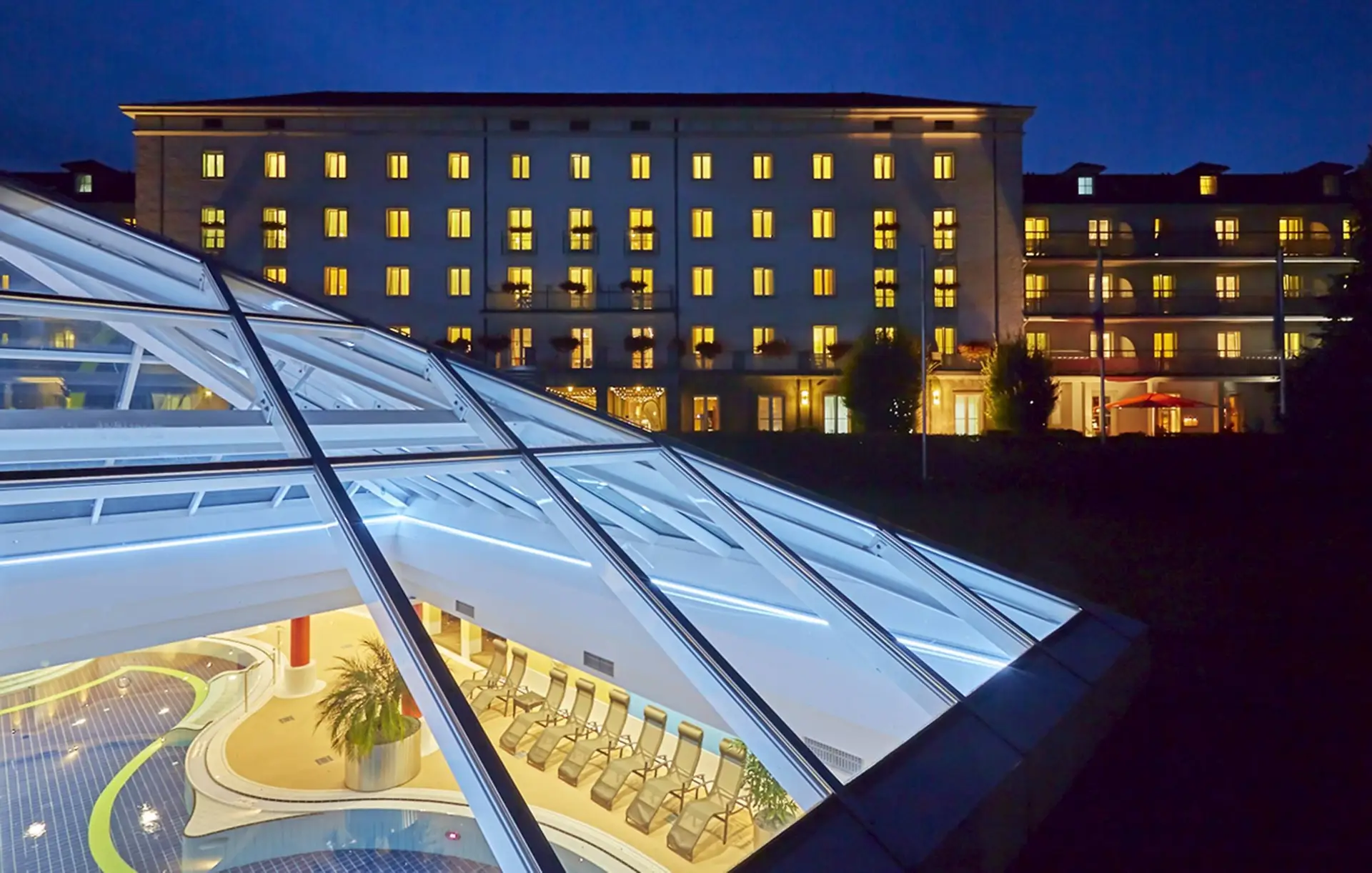 Gläsernes Dach vom Wellnessbereich beleuchtet. Im Hintergrund steht das Hotel.
