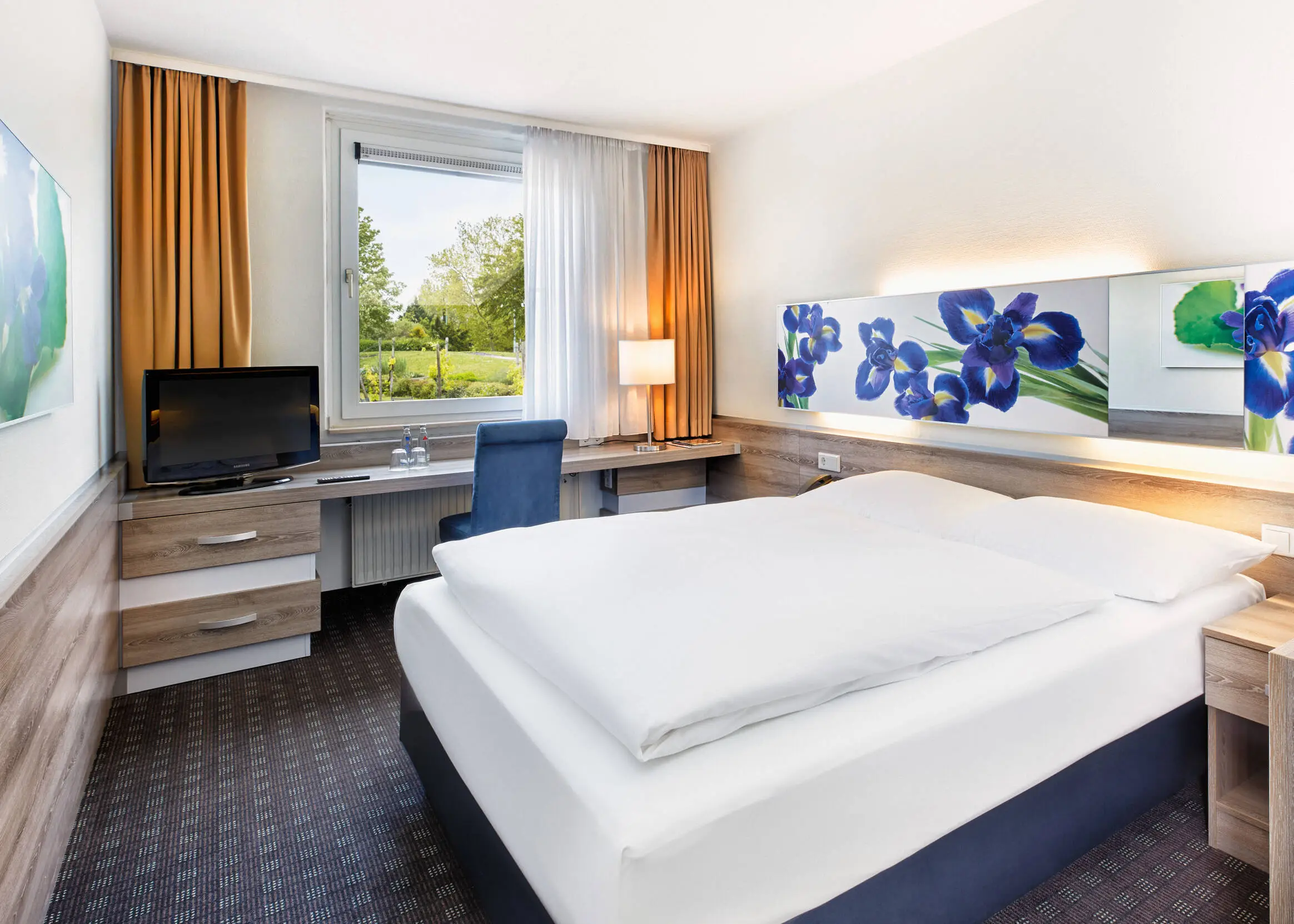 Gemütliches Hotelzimmer mit einem Bett und einem Schreibtisch daneben. Aus dem Fenster sieht man einen Garten.