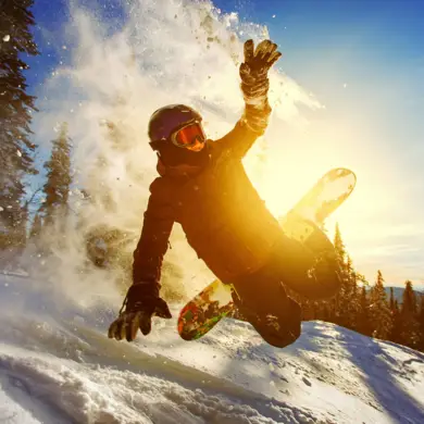 Ein Snowboarder springt und winkelt seine Beine an. Hinter ihm scheint die untergehende Sonne an ihm vorbei.