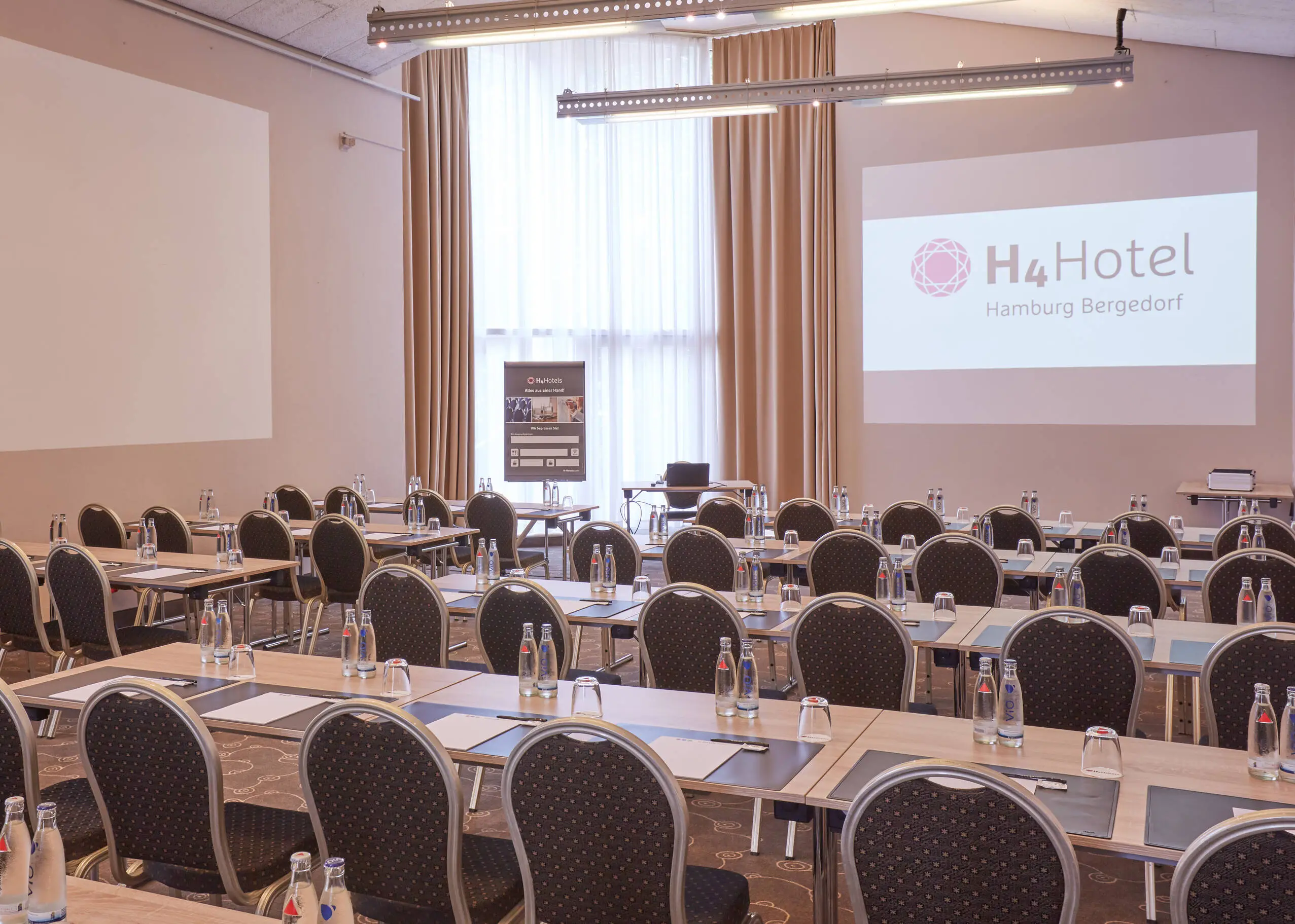 Tagungsraum mit Stuhlreihen und hohen Wänden. An die Wand wird mit einem Beamer das Hotelloge gestrahlt.