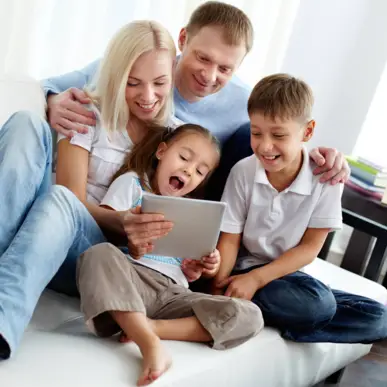 Eine vierköpfige Familie sitzt auf einer Couch und schaut freudig auf ein Tablet.