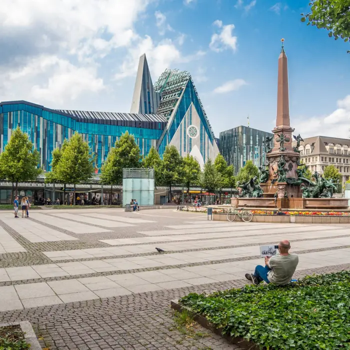 Augustusplatz mit einem großen Brunnen auf der rechten Seite. Im Vordergrund sitzt ein Mann auf Steinen und malt.