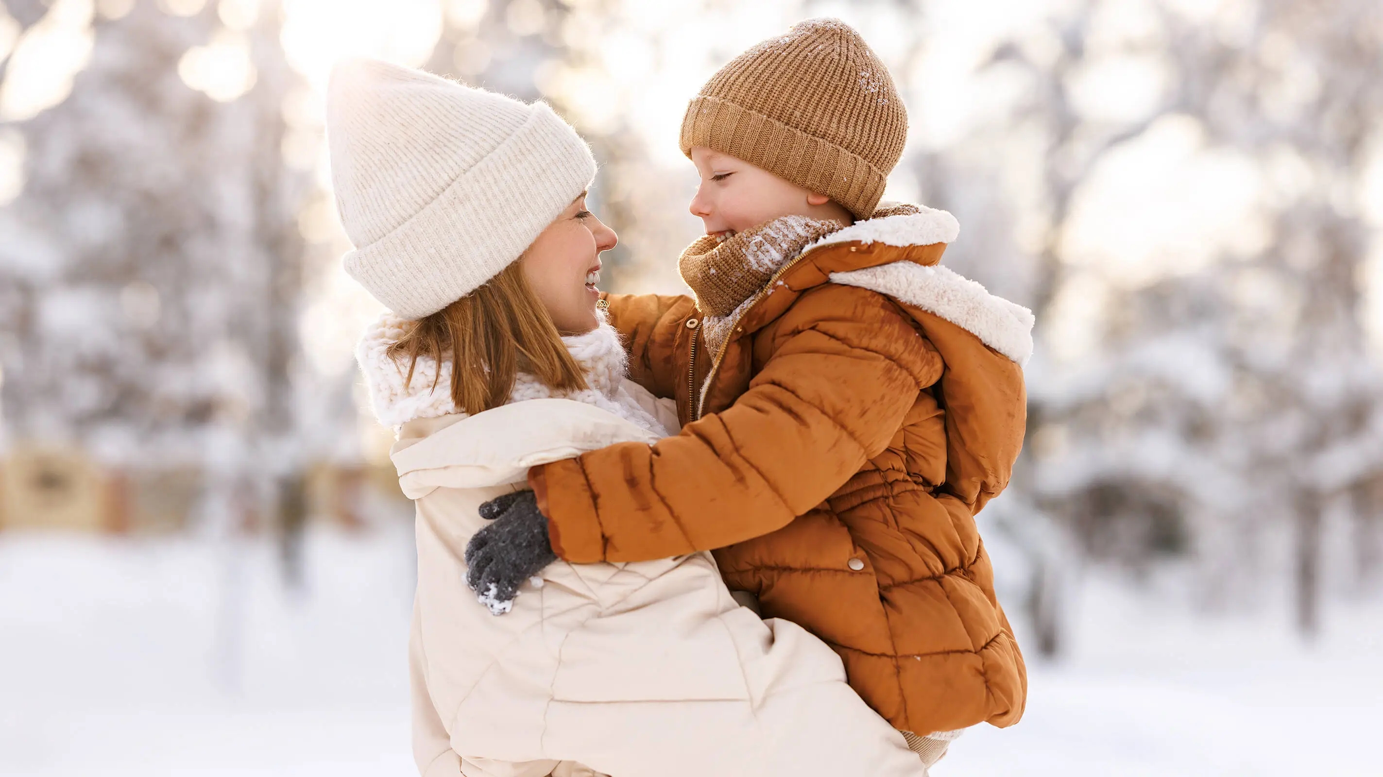 Una mujer con chaqueta y gorro blancos sostiene a un niño con chaqueta y gorro marrones. Al fondo se ve mucha nieve.