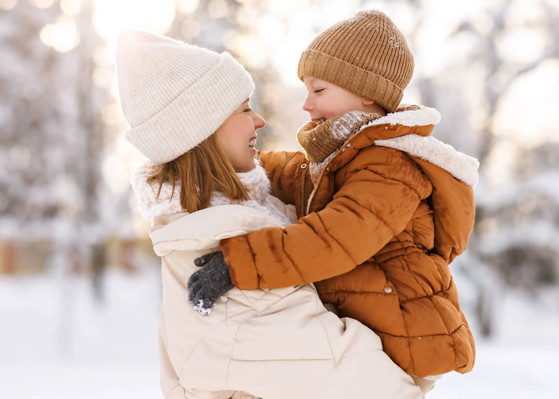 Una mujer  con chaqueta y gorro blancos sostiene a un niño con chaqueta y gorro marrones. Al fondo se ve mucha nieve.