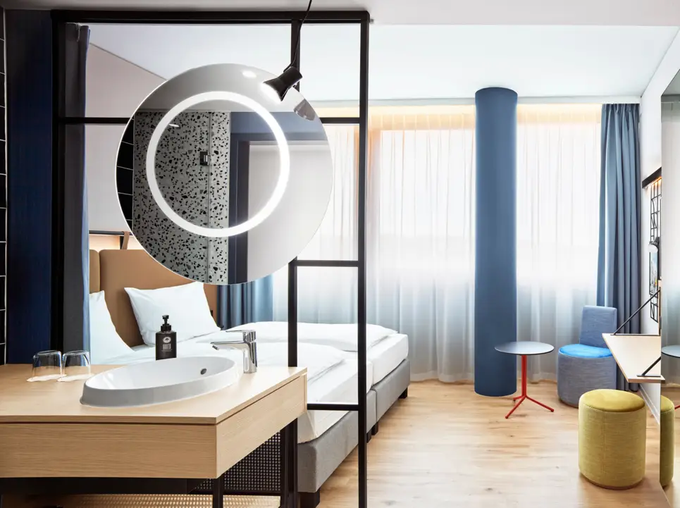 Hotelzimmer mit einem Doppelbett und einem halboffenen Badezimmer. Vor dem Bett stehen kleine Sitzmöglichkeiten.
