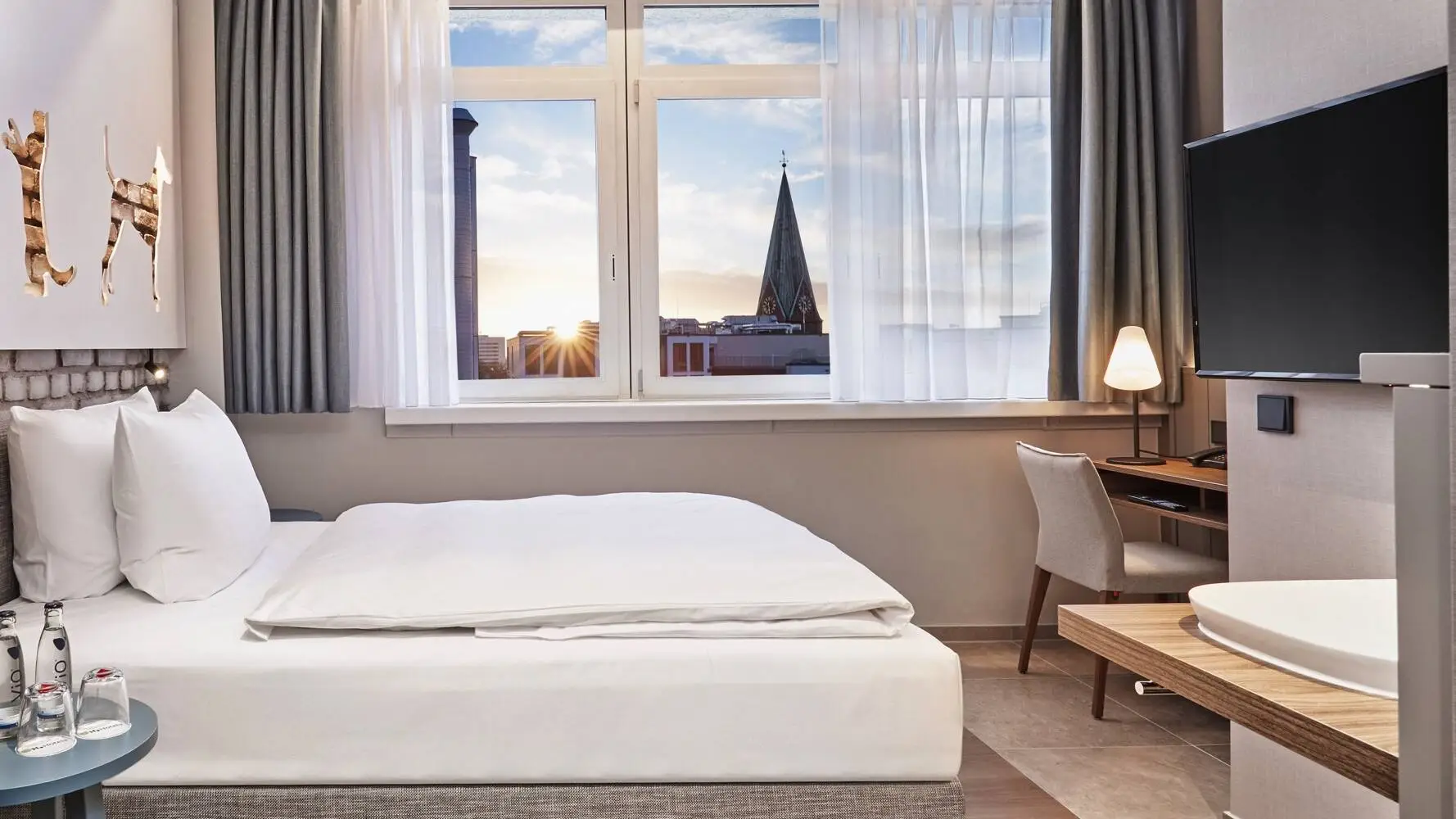 Habitación de hotel con una cama y un escritorio delante. En primer plano a la izquierda hay un armario con una cafetera y a la derecha cuelga un televisor.