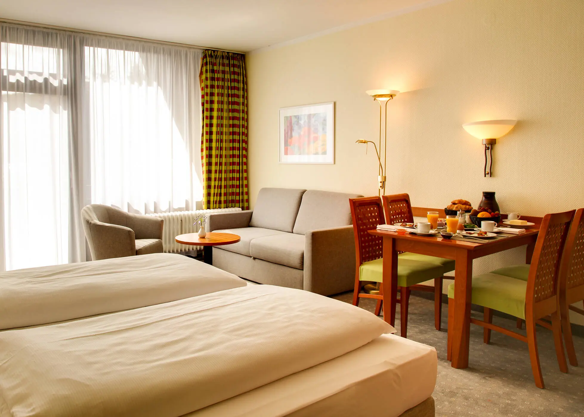 Großes Hotelzimmer mit einem Doppelbett und einem großen Fenster. Vor dem Bett steht ein Esstisch und eine kleine Sitzecke mit einer Couch.