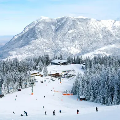 Winterlandschaft mit einem großem Berg im Hintergrund. Menchen fahren im Vordergrund eine Skipiste runter.