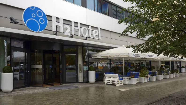 Eingang vom Hotel mit Sitzplätzen und Sonnenschirmen auf dem Gehweg daneben.
