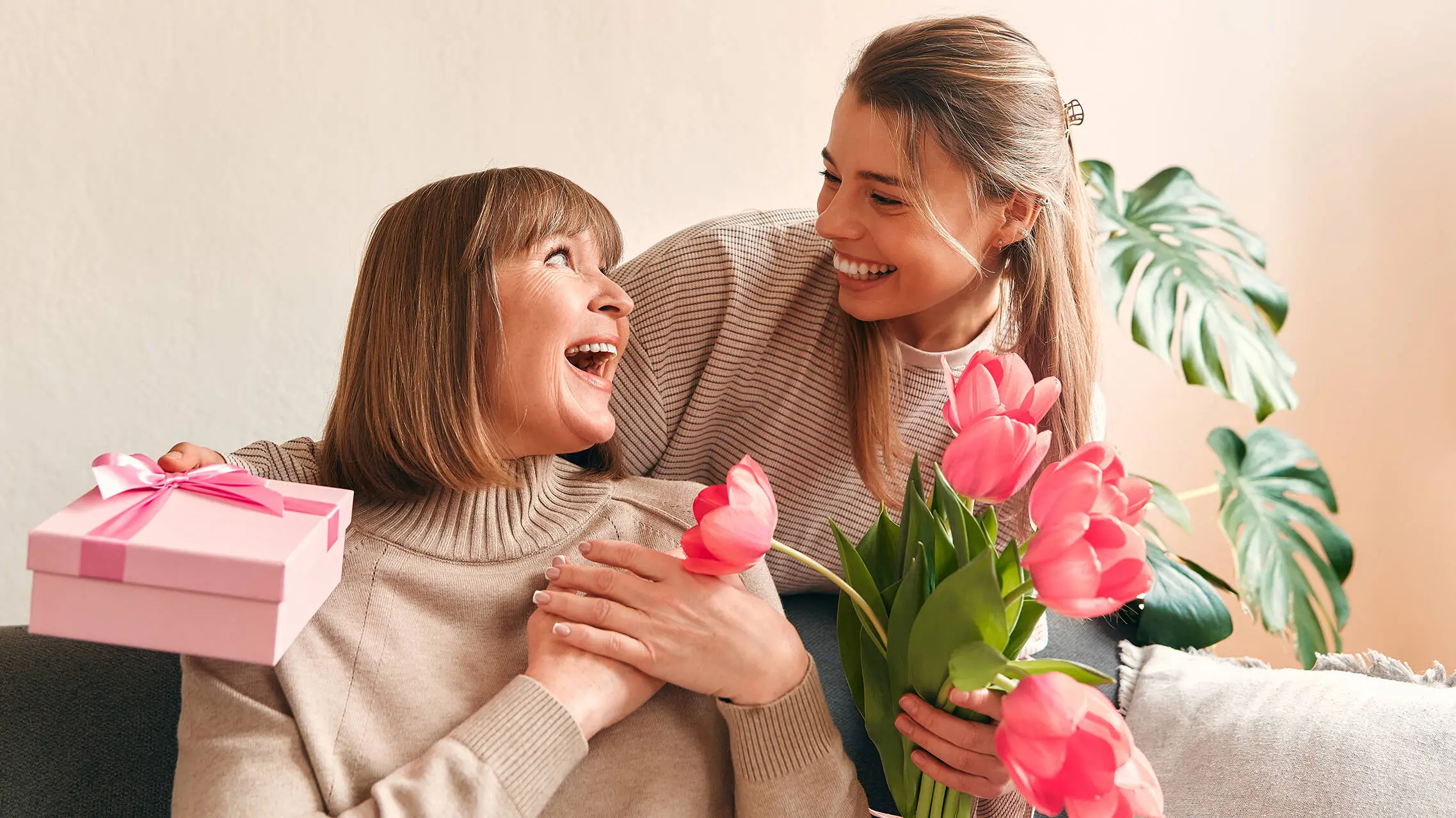 Zwei Frauen schauen sich lächelnd an. Eine Frau hat Blumen und ein Geschenk in der Hand.