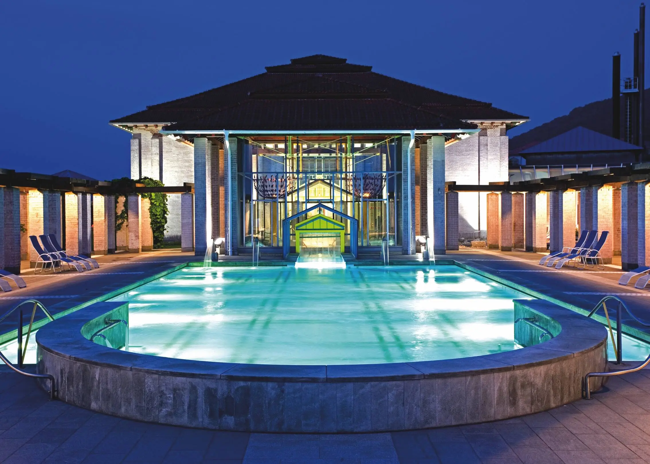 Illuminated outdoor pool at the Limes-Thermen thermal spa with sun loungers on the left and right of the terrace next to the pool.