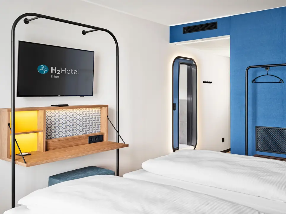 View over the bed to a wall with TV and hanging writing desk with stool.