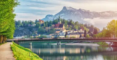 Blick über den Fluss auf eine Brücke. Auf der linken Seite ist eine kleine Promenade und im Hintergrund steht eine kleinere Kirche und Häuser.