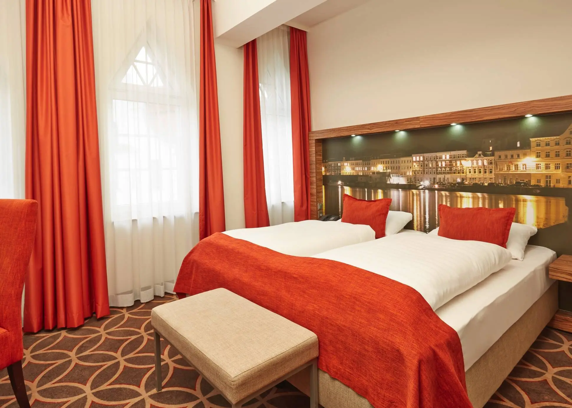 Hotelzimmer mit roten Akzenten. Hinter dem Bett hängt ein großes Bild vom Lübecker Hafen bei Nacht.