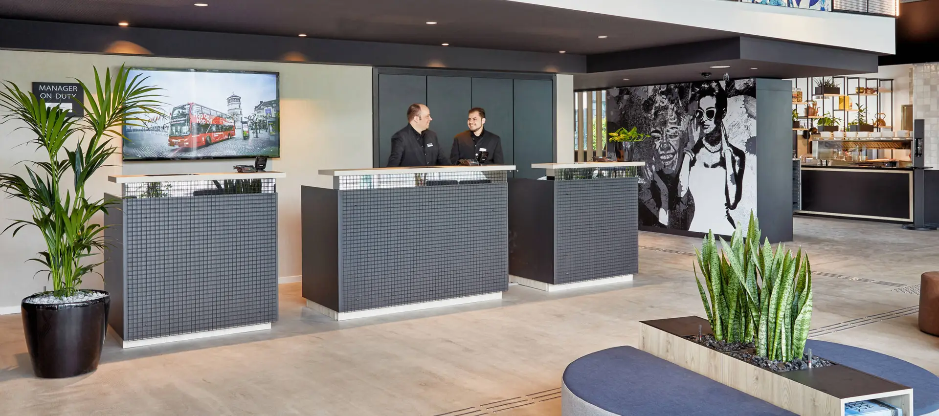 Rezeption mit drei Terminals, an der Wand steht "H2 Hotels". Vor der Rezeption steht eine Bank, die das Wort "Hello" bildet.