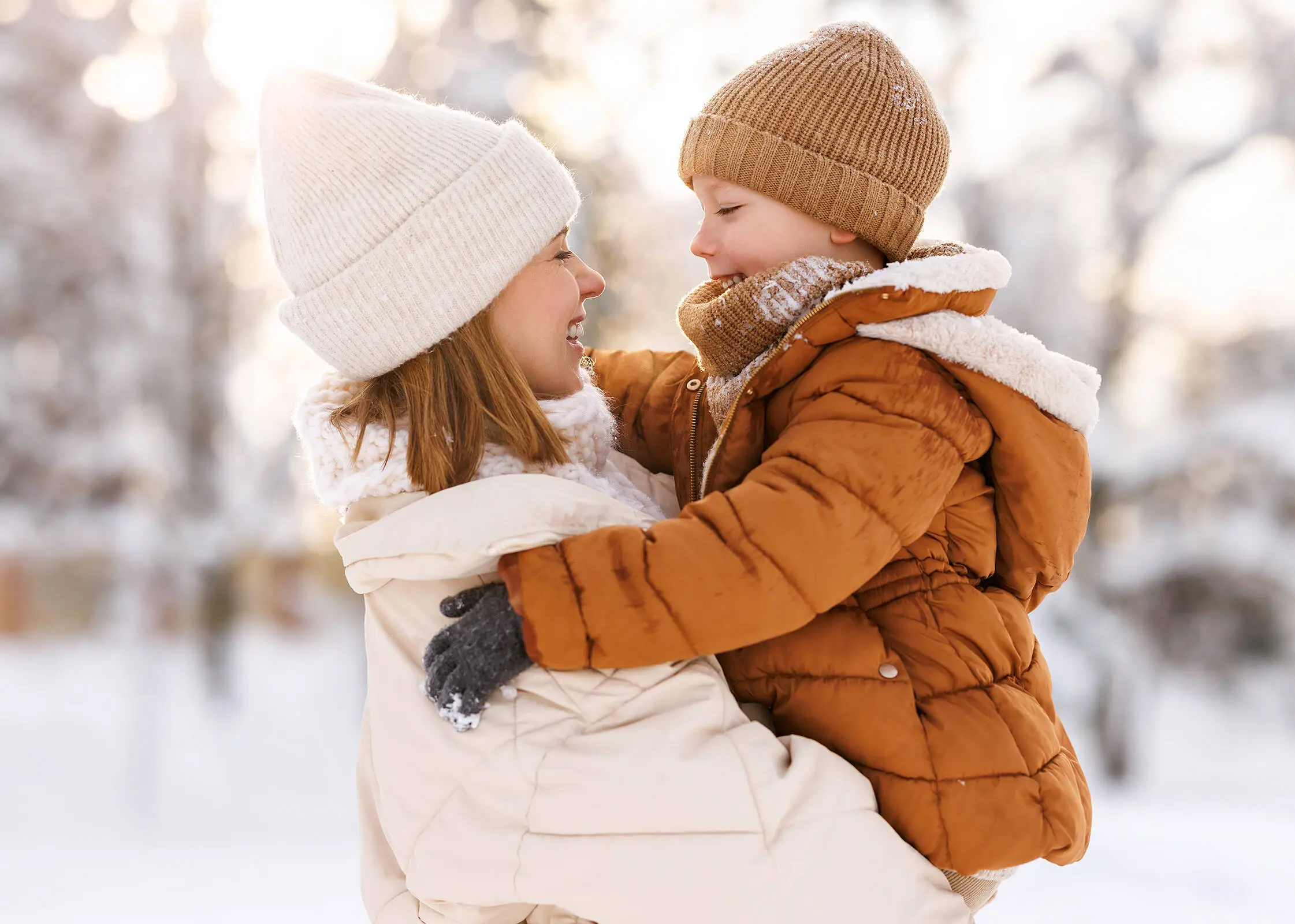 A woman wearing a white jacket and hat is holding a child wearing a brown jacket and hat. There is a lot of snow in the background.
